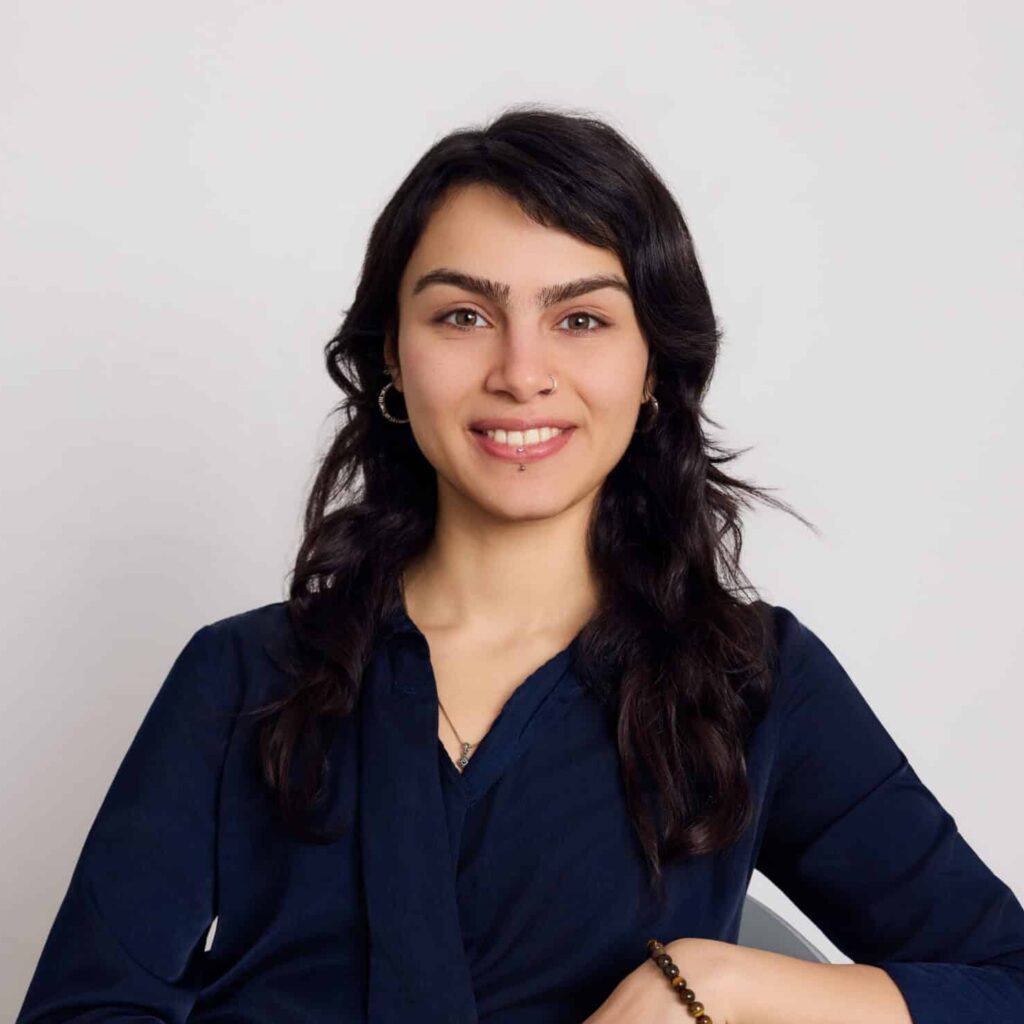 Headshot of Elnaz smiling, with shoulder-length dark hair, wearing a navy blue top against a light neutral background.